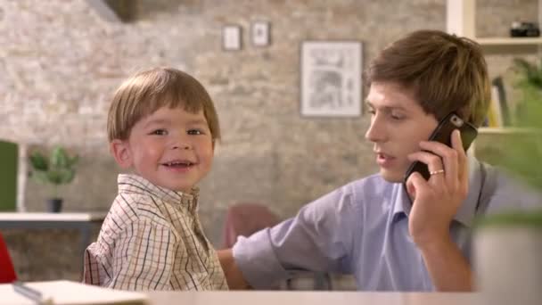 Giovane padre d'affari che gira intorno al suo piccolo figlio sulla sedia e parla al telefono in un ufficio moderno — Video Stock