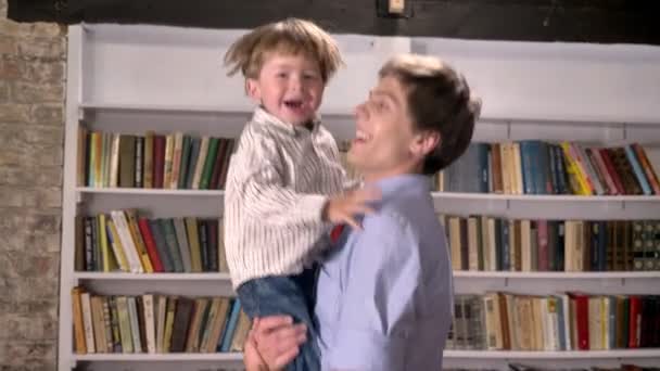Молодой отец держит своего маленького сына и прыгает в библиотеку, улыбаясь и радуясь — стоковое видео
