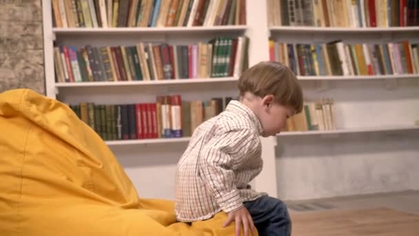 Kleine schattige jongen zittend op een stoel en het vangen van de bal van iemand, boekenkasten achtergrond — Stockvideo