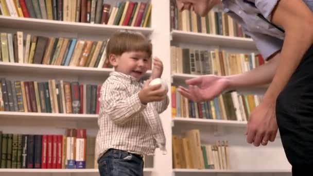 Молодой отец и его маленький сын играют с мячом, улыбаются, счастливы, полки с книжным фоном — стоковое видео