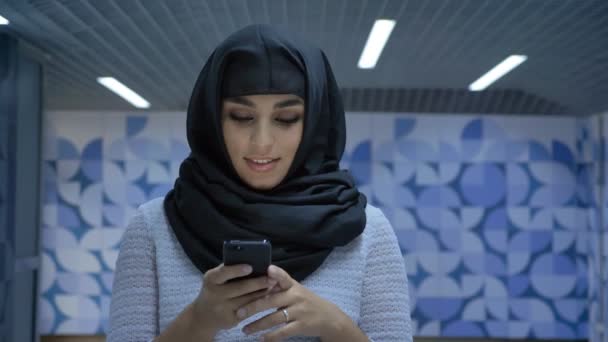 Молодая очаровательная мусульманка в хиджабе печатает сообщение на смартфоне в подземке, наблюдает за камерой, улыбается, религия концепция, коммуникация — стоковое видео