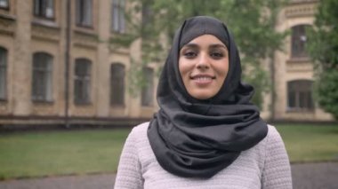 Hijab genç güzel Müslüman kızı ayakta ve gündüz, kötü havada gülümseyerek kameraya, arka planda, religiuos kavram bina izliyor