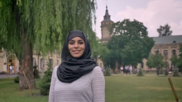 Joven chica musulmana dulce en hijab está de pie y sonriendo durante el día, con mal tiempo, mirando a la cámara, construyendo sobre el fondo, concepto religiuos — Vídeos de Stock