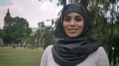 Genç Müslüman kız hijab içinde gündüzleri kötü havada Park kameraya, arka planda, religiuos kavram bina izlerken gülümsüyor