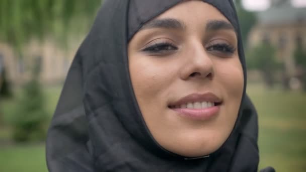 Gezicht van jonge mooie moslim meisje in hijab is permanent en glimlachend overdag, bij slecht weer, kijken in de camera, religiuos concept, onscherpe achtergrond — Stockvideo