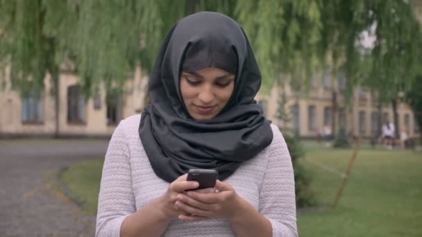 年轻美丽的穆斯林女孩在面纱是在她的智能手机上键入信息, 看着相机和微笑在蓝色的天气, 沟通概念, 宗教概念 — 图库视频影像