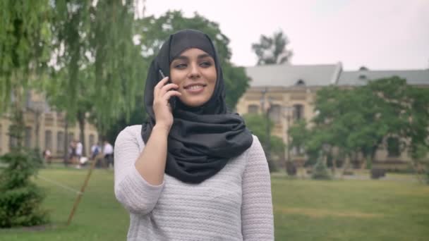 Menina muçulmana jovem no hijab está falando no telefone em tempo azul no parque, construindo em segundo plano, conceito religiuos, conceito de comunicação — Vídeo de Stock