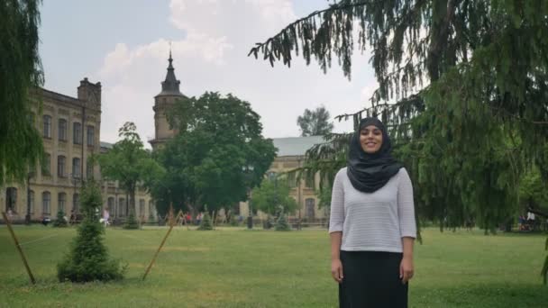 Jovem bela garota muçulmana no hijab está de pé e sorrindo durante o dia, desfrutando do tempo, construindo no fundo, conceito religiuos — Vídeo de Stock