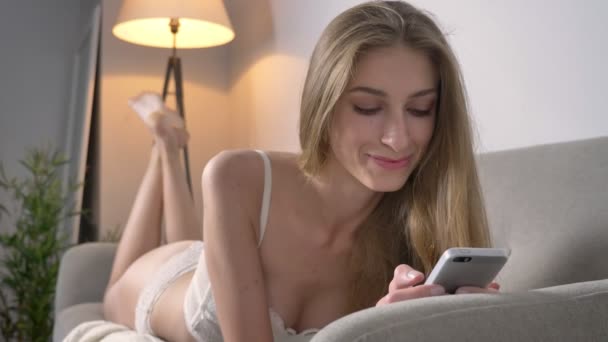 Mladá blond ženy v prádlo ležet na gauči a chatování na telefonu, s úsměvem — Stock video