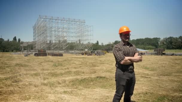 Självsäker builder stående i hjälm med korsade händer, metall konstruktion på bakgrunden, halm fält — Stockvideo