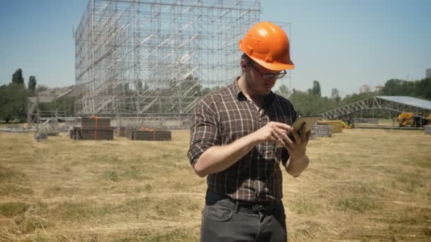 Architetto in casco e occhiali con tablet e in piedi su campo di paglia, costruzione in metallo su sfondo, concentrato e serio — Video Stock