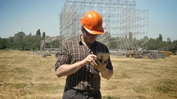 Fiatal builder sisak állandó tabletta a szalma mező, fém telepítés a háttérben, koncentrált — Stock videók