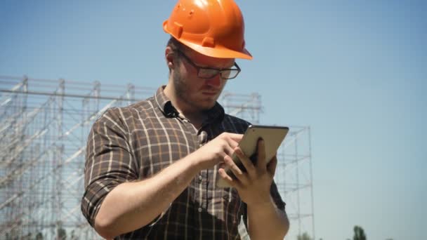 Arquitecto joven usando tableta y frunciendo el ceño, construcción de metal sobre fondo, serio — Vídeos de Stock