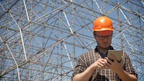 Joven arquitecto en proyecto de comprobación de casco en tableta y fruncir el ceño, construcción de metal sobre fondo, serio y preocupado — Vídeos de Stock