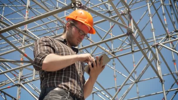 Arquitecto joven en casco usando tableta y mirando la instalación de metal en el fondo, la planificación y el diseño de un nuevo proyecto, preocupado — Vídeos de Stock