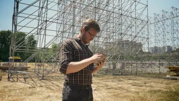 Hombre joven en gafas usando tableta y mirando la construcción de metal en el fondo, de pie en el campo de paja, serio y preocupado — Vídeos de Stock