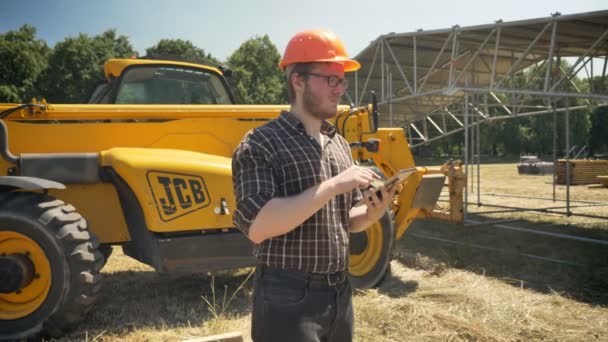 Młody budowniczy w okularach za pomocą tabletu i marszcząc brwi, patrząc w przyszłość, stojąc na polu słomy, ciągnik na tle — Wideo stockowe