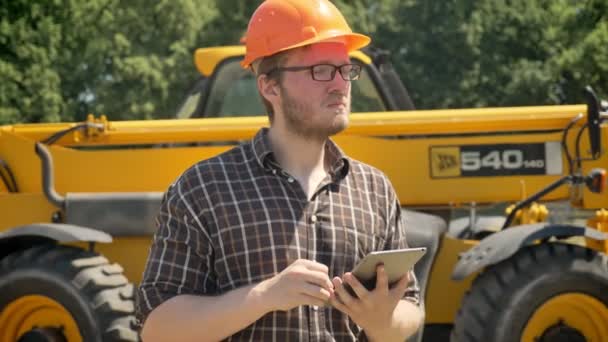 Porträt eines jungen Bauarbeiters mit Helm, Tablet in der Hand und Stirnrunzeln, nach vorne blickend, nachdenklich, Traktor im Hintergrund — Stockvideo