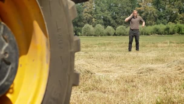 Joven en gafas hablando por teléfono y caminando en el campo de paja, preocupado, detrás del tractor, campo — Vídeo de stock