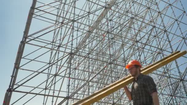 Joven constructor en casco que lleva tablón de metal pesado, trabajando cerca de una instalación enorme — Vídeos de Stock