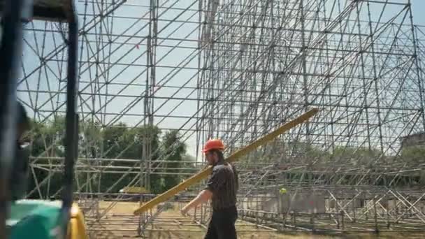 Молодой строитель в шлеме, несущий тяжелые металлические доски, работает рядом с огромной установкой на соломенном поле — стоковое видео