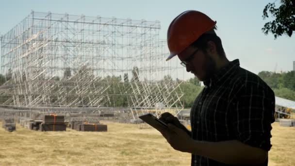 Νέος αρχιτέκτονας στο κράνος, εξετάζοντας νέα μεταλλική κατασκευή και χρήση tablet, στέκεται στο άχυρο πεδίο — Αρχείο Βίντεο