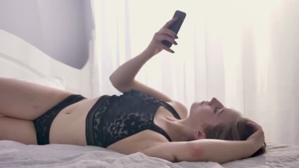 Menina sexy nova está deitada em lingerie na cama, rolando e conversando no smartphone, sorrindo, flertar conceito, conceito de comunicação — Vídeo de Stock