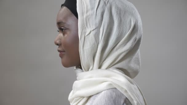 Chica musulmana africana joven en hijab está girando la cabeza y mirando a la cámara, sonriendo, concepto religioso, fondo gris — Vídeo de stock