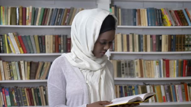 Young african muslim girl in hijab is holding book, watching at camera, religioun concept, booksheves on background — Stock Video