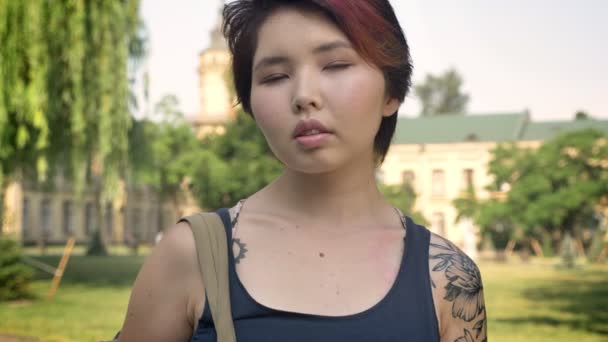 Portrait of young asian female student with tattoo standing with backpack and looking in camera in park near university, confest — Stok Video