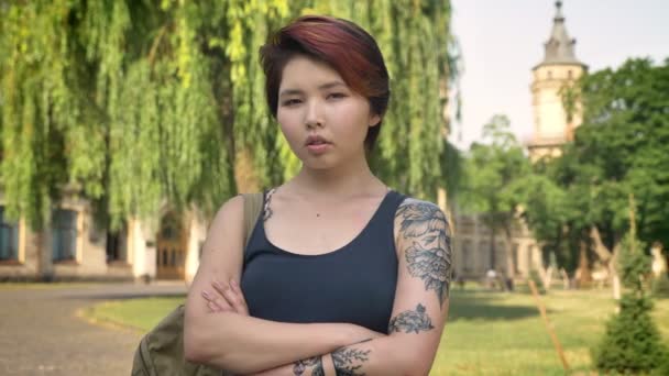 Fiatal ázsiai nők, a tetoválás állandó, keresztezett kézzel, és keresi a kamera park Egyetem közelében, magabiztos és súlyos portréja — Stock videók