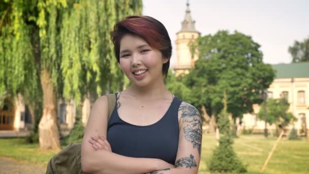 タトゥーの交差させた手に立ち、幸せな笑みを浮かべて、大学近くの公園でカメラを探している若いアジア女性の肖像画 — ストック動画