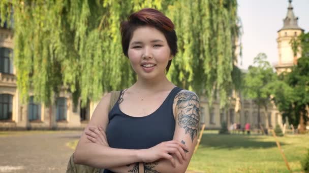 Asiatisk ung tatuerade kvinnlig student står i park, nära universitetet och tittar i kameran, leende och glad — Stockvideo