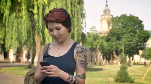 アジア系の若い入れ墨立っている女性 大学近くの公園での携帯電話のテキスト メッセージ 幸せな笑みを浮かべて — ストック動画