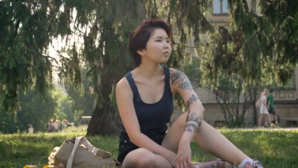 Trendiga unga asiatiska kvinnlig hipster med tatuering sitter på gräset i parken nära university, tankeväckande — Stockvideo