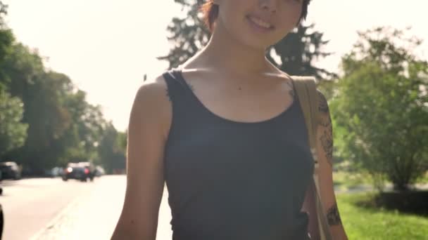 Giovani donne asiatiche con mano tatuata che tiene lo skateboard e lo zaino, camminando in avanti nel parco vicino all'università — Video Stock