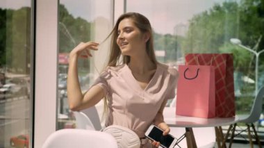 Genç güzel kız kafede büyük pencereler gündüz de dokunmadan saç, holding smartphone, çanta masada oturuyor