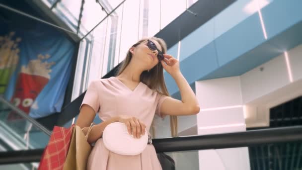 Joven chica atractiva está de pie en el centro comercial, se quita las gafas de sol, llevar bolsas, concepto de compras, concepto de moda — Vídeos de Stock