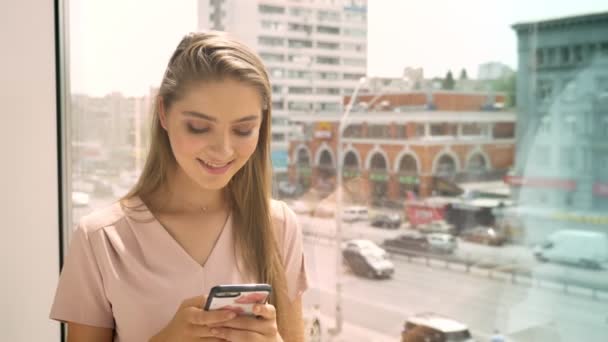 Joven hermosa chica está sentado en la ventana, chating en el teléfono inteligente en el día, concepto de comunicación, concepto urbano — Vídeos de Stock