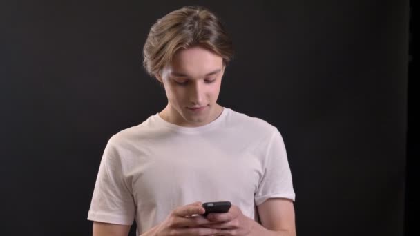 Joven hombre guapo en camisa blanca escribiendo en el teléfono y mirando a la cámara, aislado sobre fondo negro — Vídeos de Stock