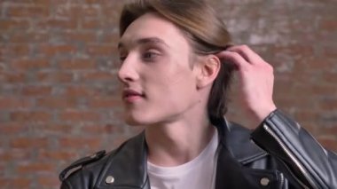 Saçları çok dokunaklı ve tuğla arka plan üzerinde izole kamera, gülümseyen siyah deri ceketli genç yakışıklı adamı