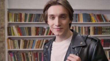 Kütüphanede duran ve seyir fotoğraf makinesi, gülümseyen, kitap raf arka plan siyah deri ceketli genç yakışıklı adam