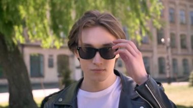 Gözlüklerini yetiştirme ve kamera, kentsel sokakta duran bakarak siyah deri ceketli genç yakışıklı adamı