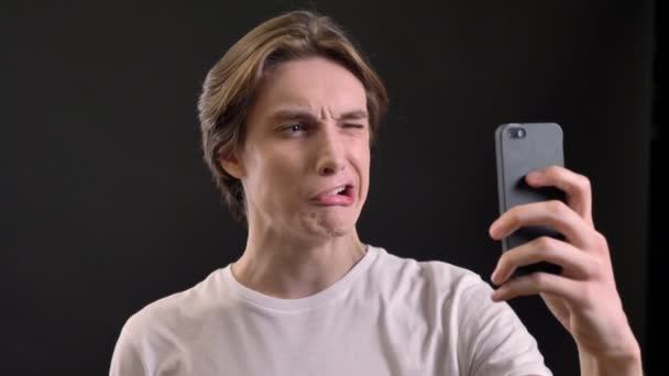 Όμορφος νεαρός λήψη selfie και κάνοντας διάφορα αστεία πρόσωπα, απομονώνονται σε μαύρο φόντο — Αρχείο Βίντεο