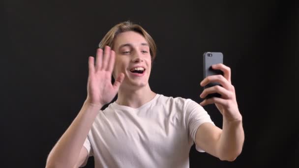 Jovem homem bonito em camisa branca fazendo videochamada no smartphone, sorrindo, isolado sobre fundo preto — Vídeo de Stock