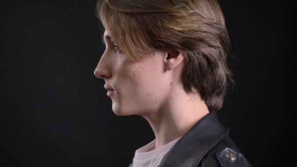 Young charming man in black leather jacket standing in profile, turning and looking at camera, isolated over black background, side profile view — Stock Video