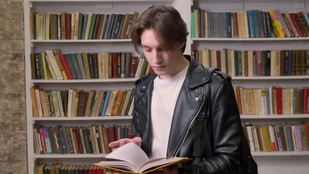 Young handsome student standing in library and holding book, smiling at camera, book shelves background — Stock Video