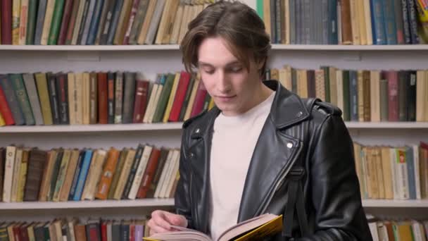 Joven hombre guapo en chaqueta de cuero negro libro de lectura en la biblioteca, sonriente, retrato de estudiante masculino estudiando — Vídeo de stock