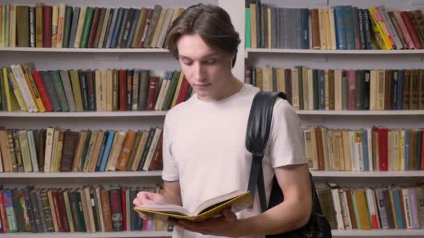 Young beautiful man in white shirt standing in library and reading, looking at camera and holding book, bookshelves background, serious — Stock Video