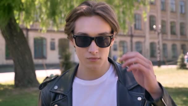 Portrait of young handsome man in black leather jacket taking off glasses and looking at camera, street background — Stock Video